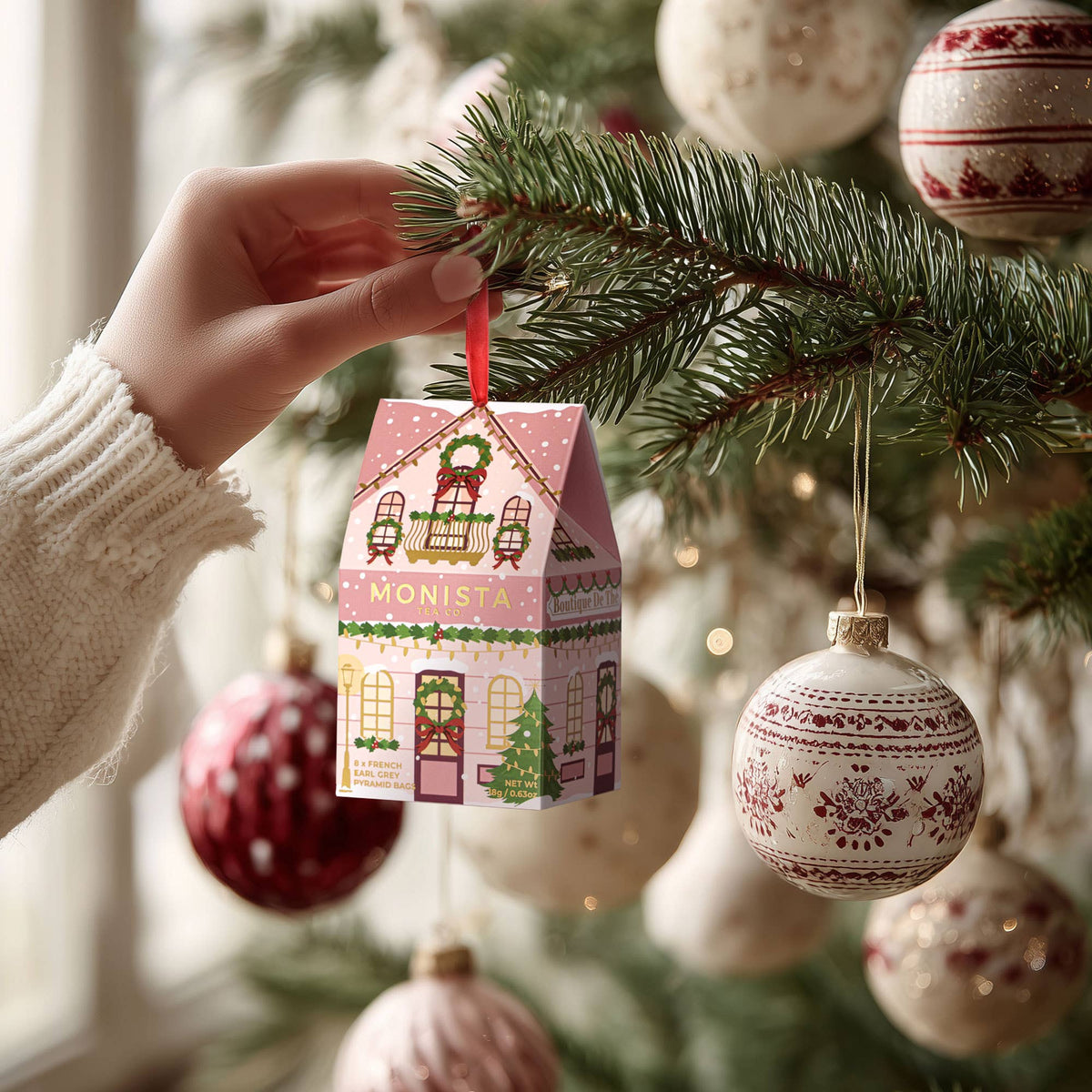 A hand wearing a cream knit sweater hangs a small pink house-shaped Christmas ornament from a pine tree branch. The ornament has a red ribbon loop and festive printed details, with other decorated ornaments blurred in the background.