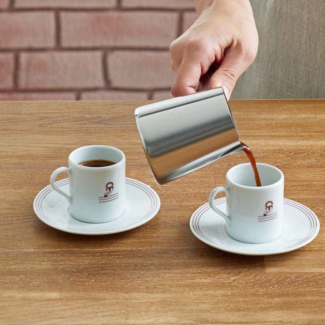 Person pouring coffee from a stainless steel pitcher into two white cups on a wooden table.