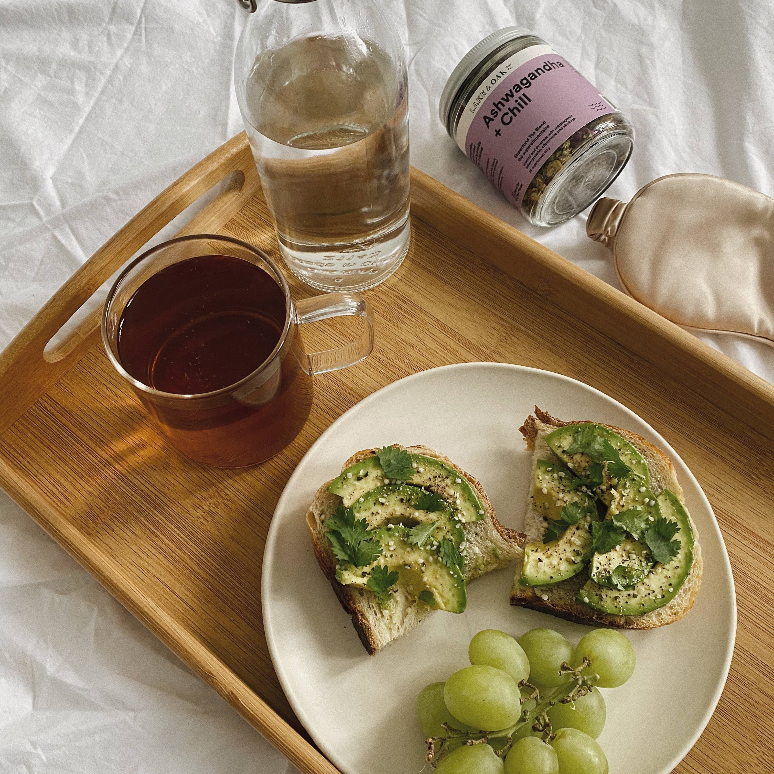 Wooden tray with a plate of toast, grapes, a glass of water, and a jar on a white surface.