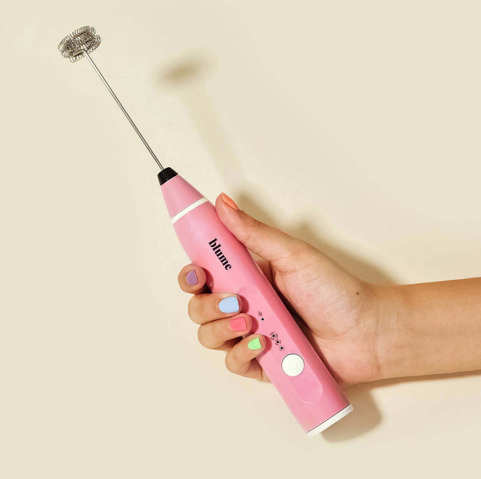 Pink handheld milk frother with whisk attachment held by a hand on a beige background