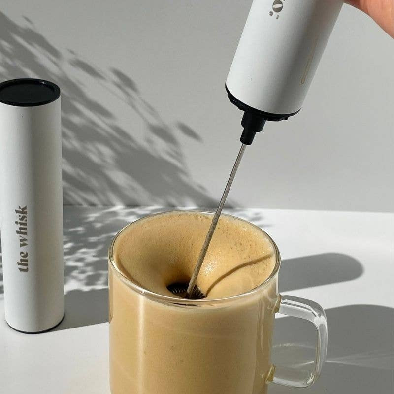Person using a milk frother to create foam in a glass of coffee on a light background