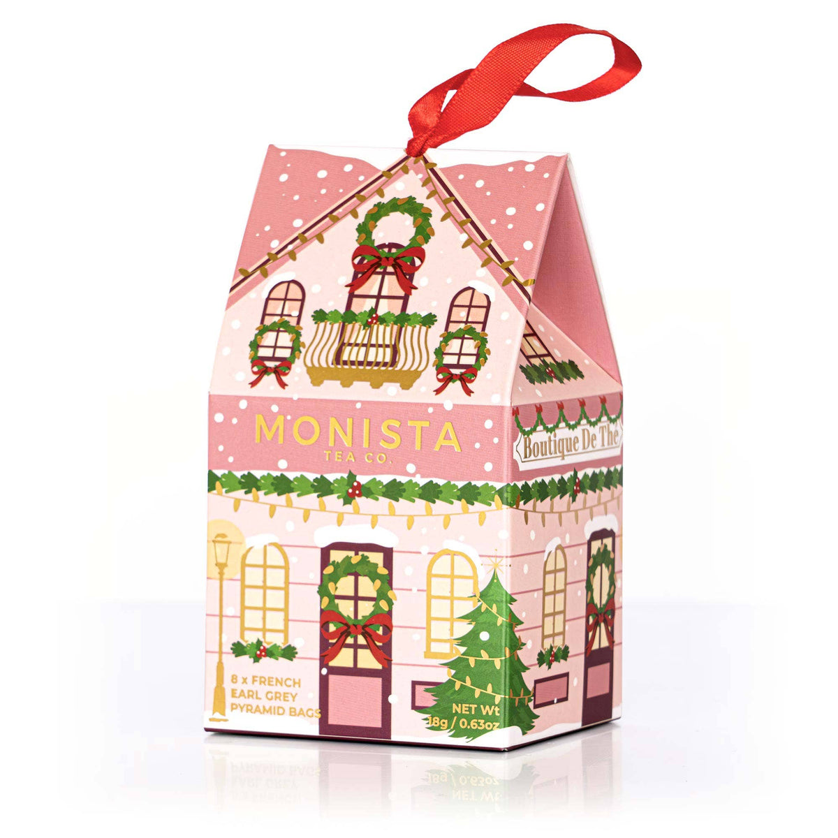 A pink house-shaped Monista Christmas tea box with a red hanging ribbon, decorated with Christmas illustrations including wreaths, lights, and a tree, on a white background.