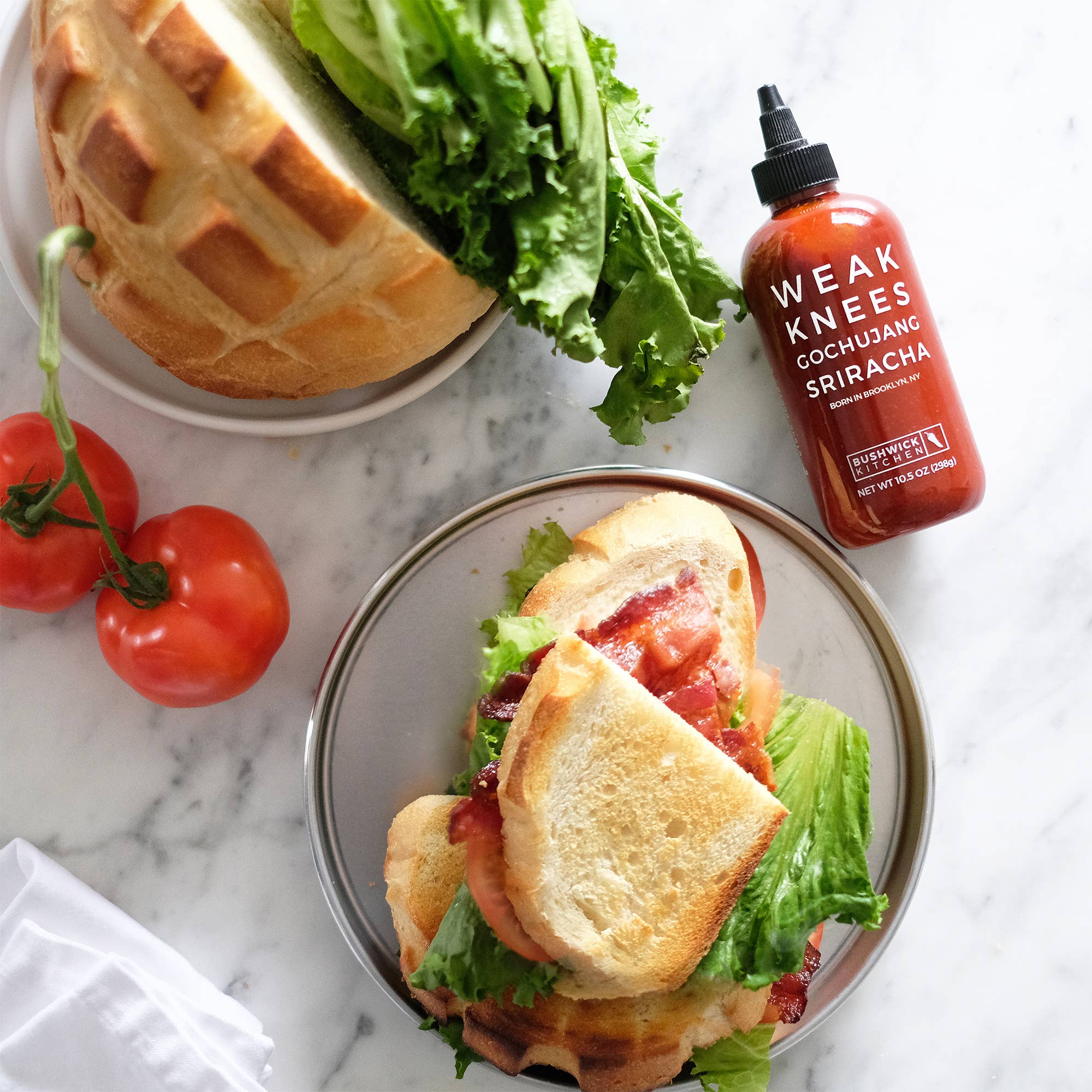 Two sandwiches with tomatoes and lettuce, a bottle of Weak Knees Sriracha sauce on a marble surface.