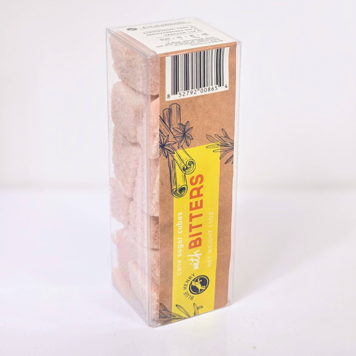 A clear plastic box of bitters sugar cubes containing pale pink sugar cubes, with a brown kraft paper label featuring a bright yellow vertical banner and botanical illustrations.