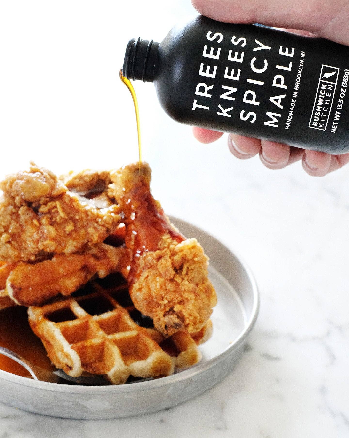 Fried chicken and waffles with a bottle of Trees Knees Spicy Maple syrup being poured over them.