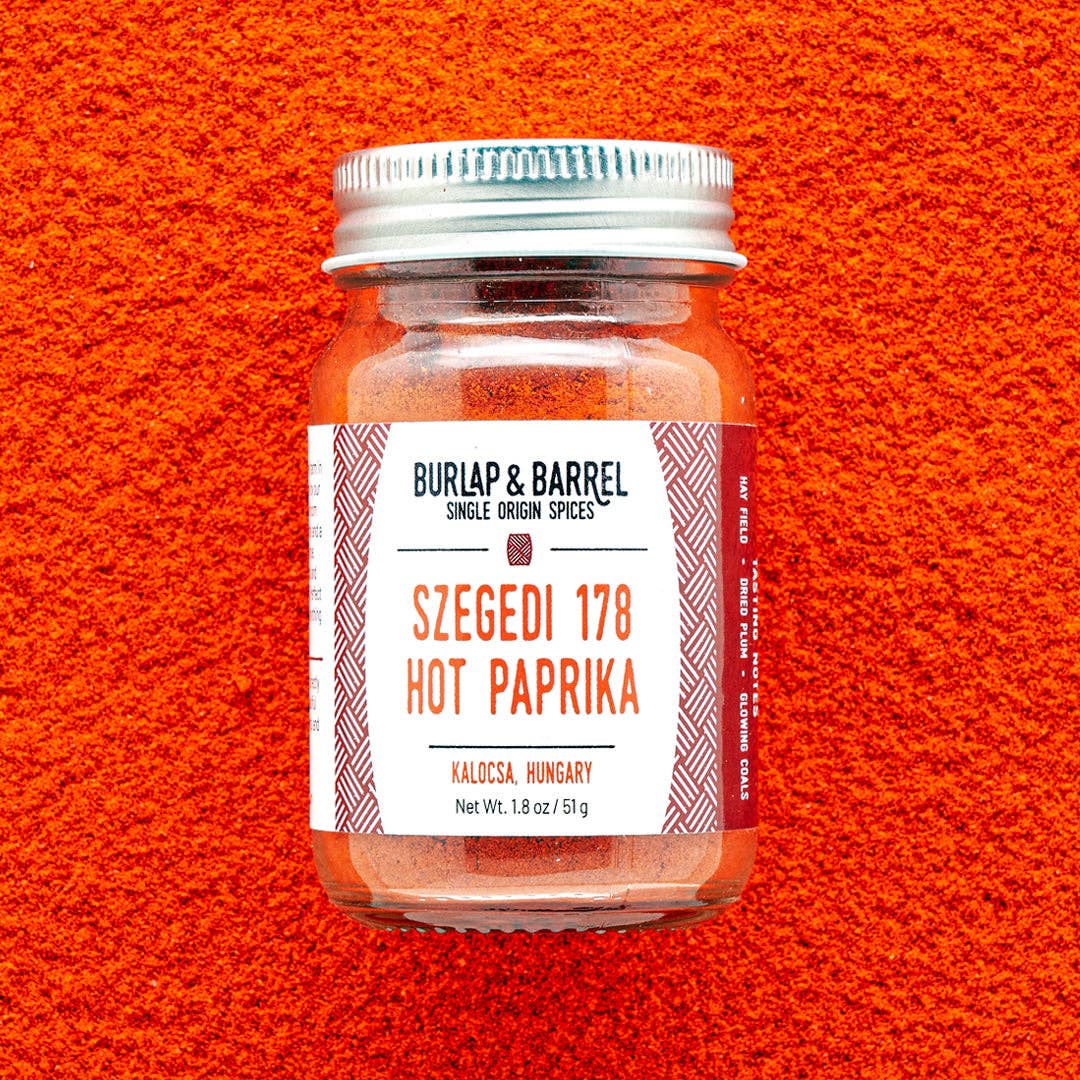 A glass jar of Burlap & Barrel Szegedi 178 Hot Paprika spices, featuring a white label with red text, set against a vibrant textured background of the spice.