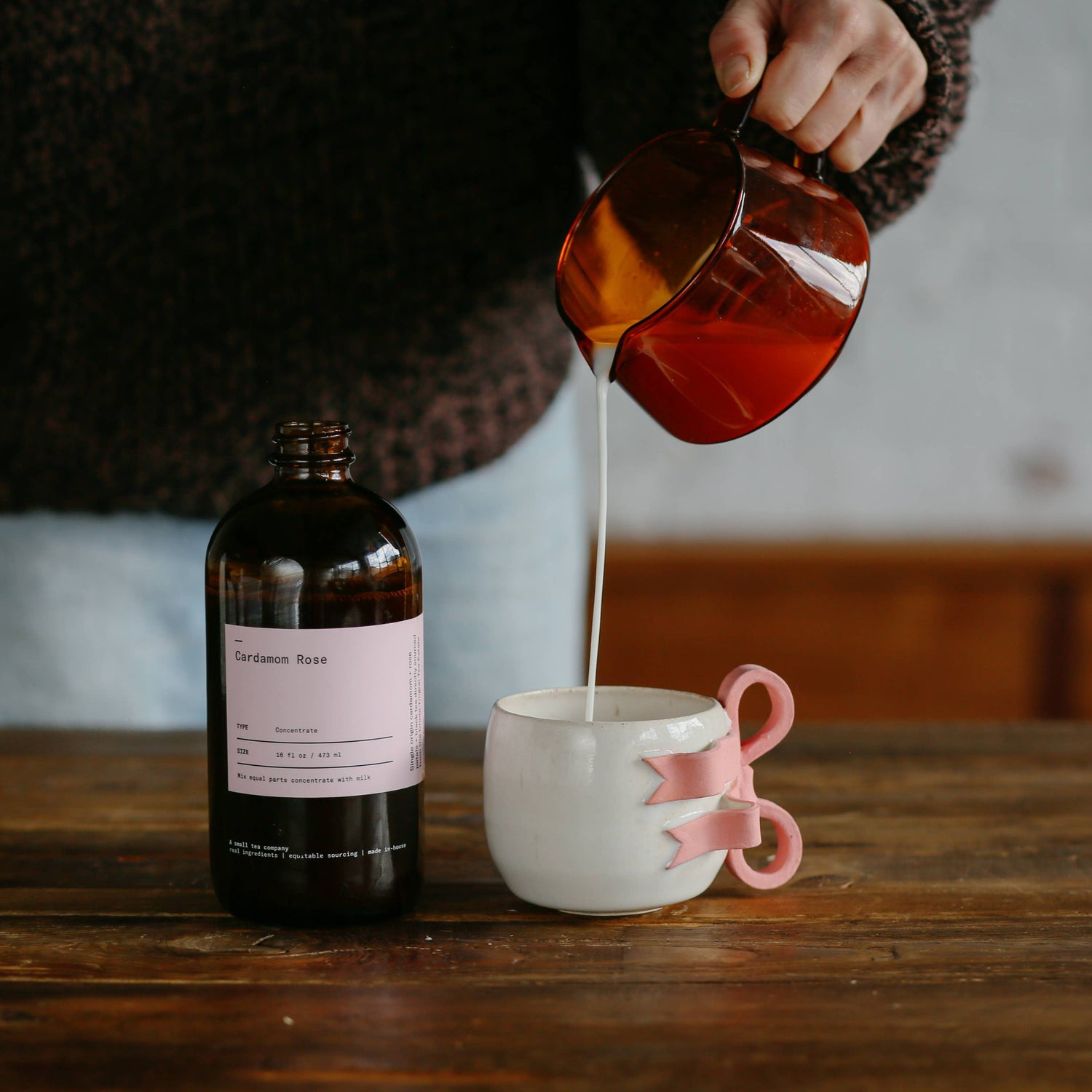 Person pouring liquid from a bottle into a mug on a wooden table.