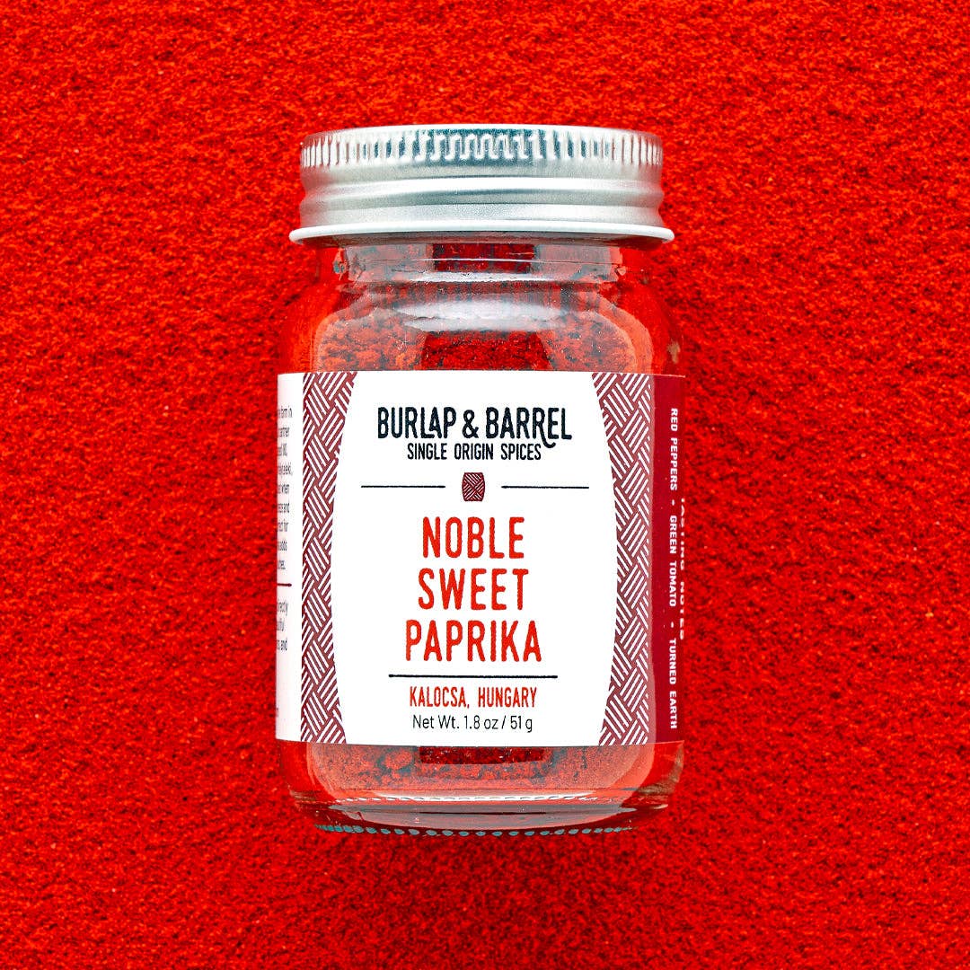 A glass jar of Burlap & Barrel Noble Sweet Paprika centered against a bright red textured background. The jar has a silver metal lid and a white label with red text and geometric patterns.