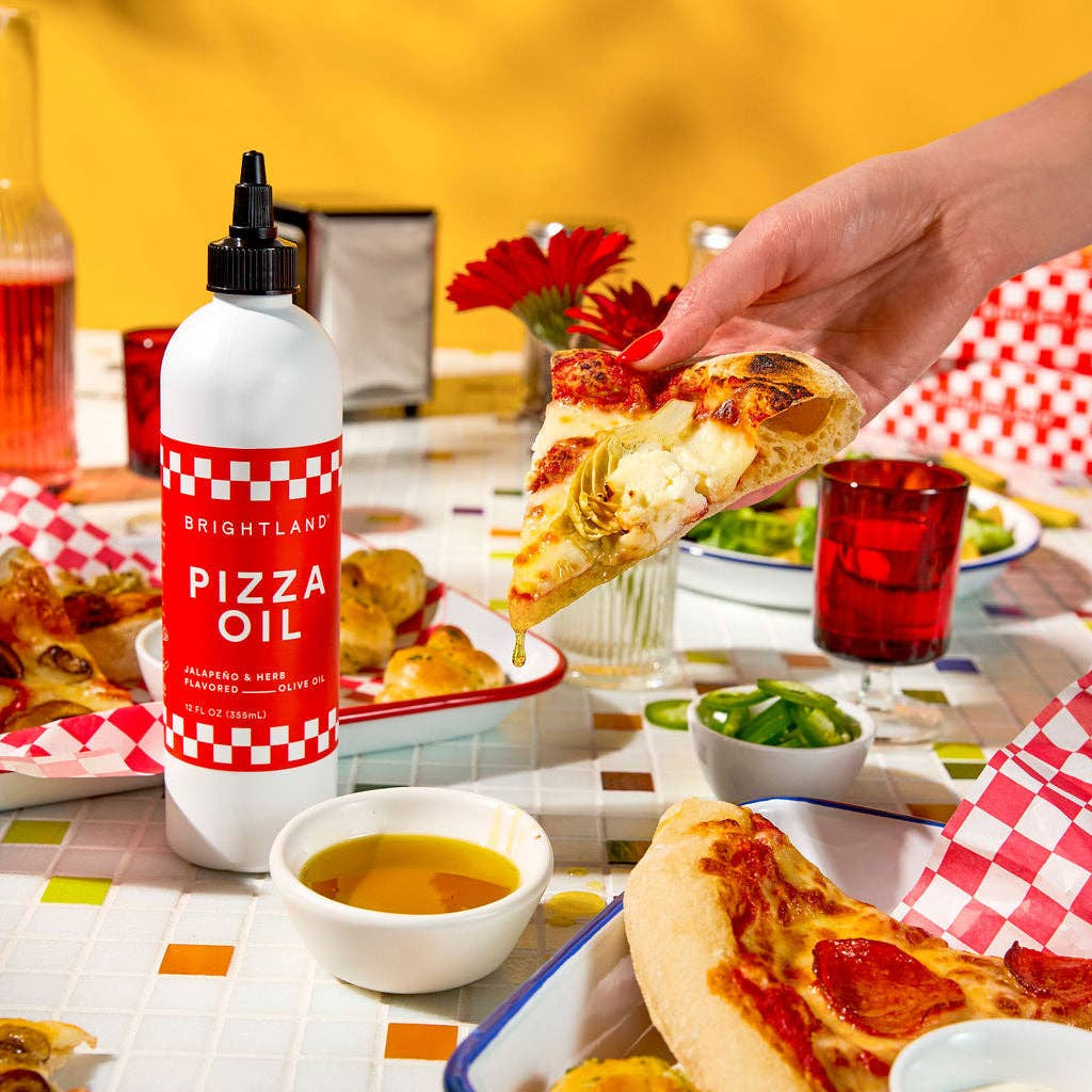 Person holding pizza slice with Brightleaf Pizza Oil bottle on a table with pizza and drinks.
