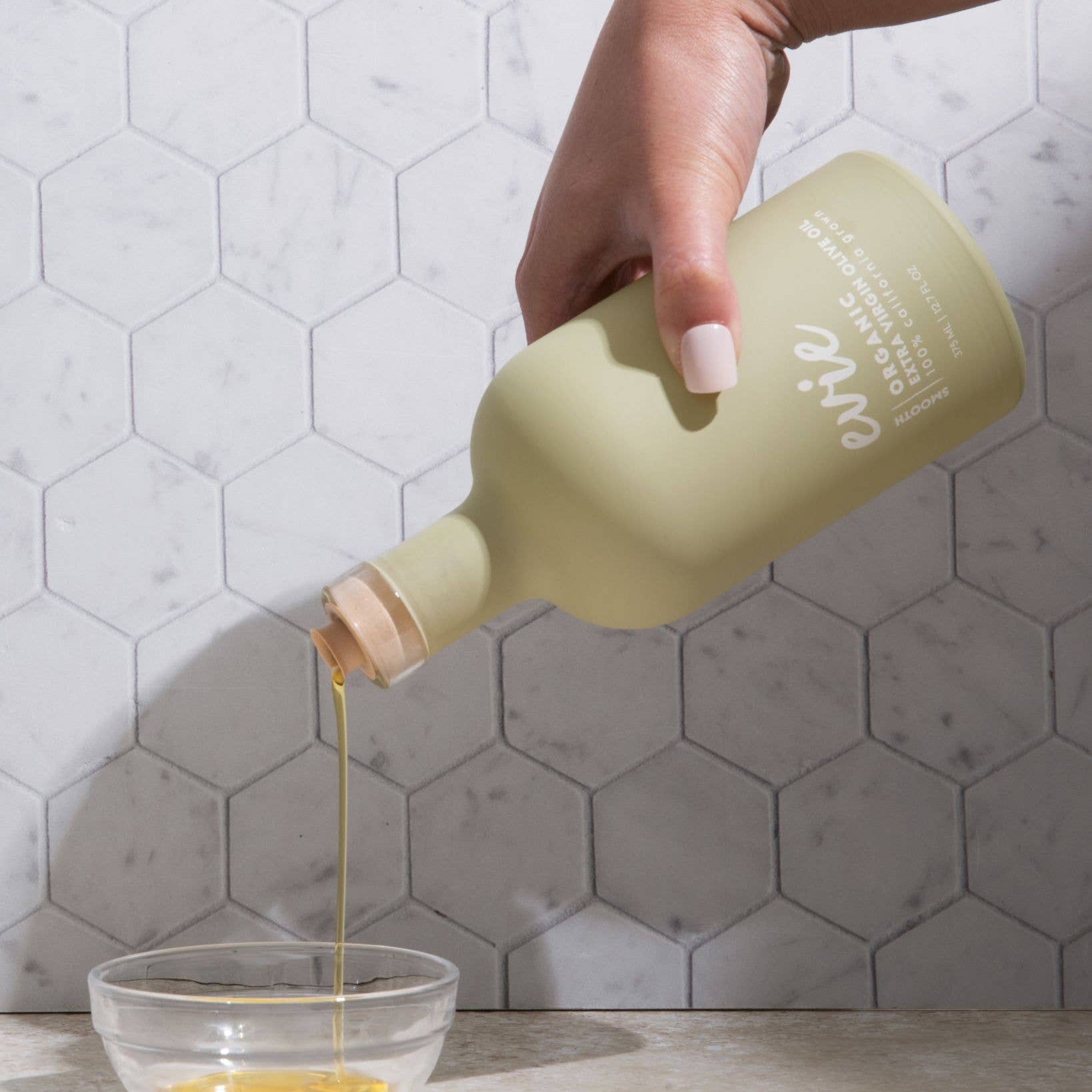Hand pouring a liquid from a green bottle into a glass bowl against a tiled wall background
