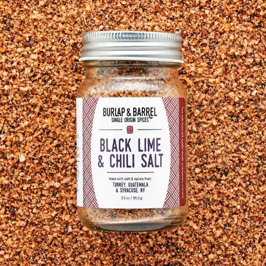 A glass jar of Burlap & Barrel Black Lime & Chili Salt, filled with coarse brown and reddish-brown spices, set against a background of the same spice blend. The jar has a silver metal lid and a white label with dark purple text and red patterned borders.