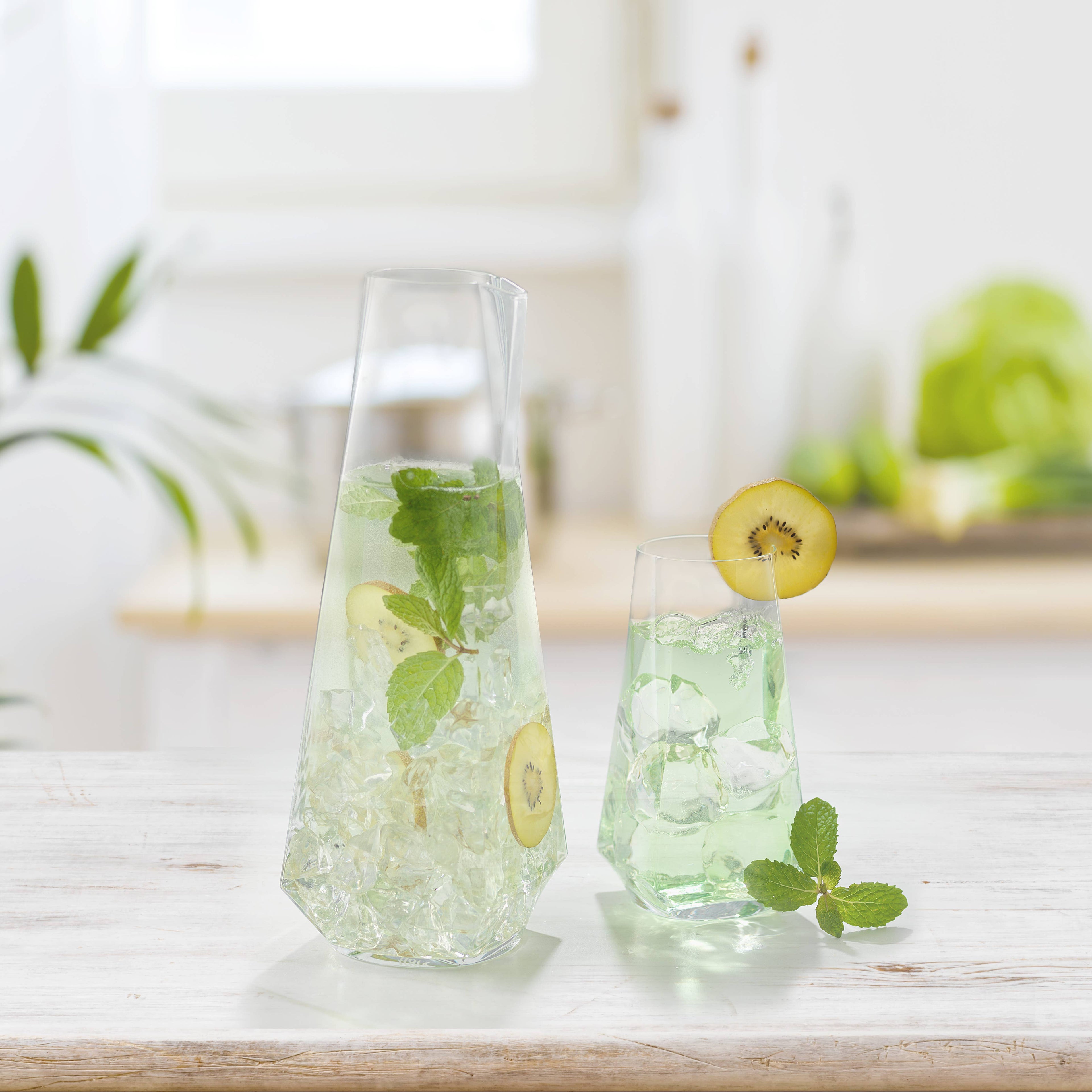 Clear carafe and glass with green liquid, kiwi slices, and mint leaves on a light surface.