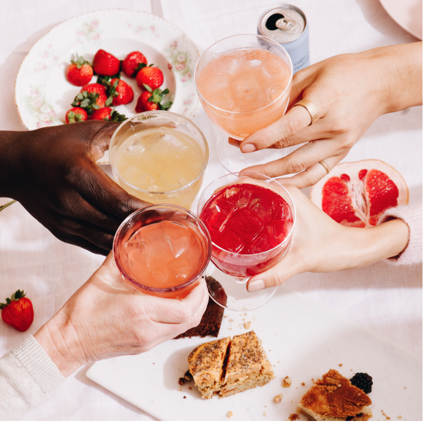People holding glasses of colorful drinks with a plate of strawberries and pastries on a white surface.