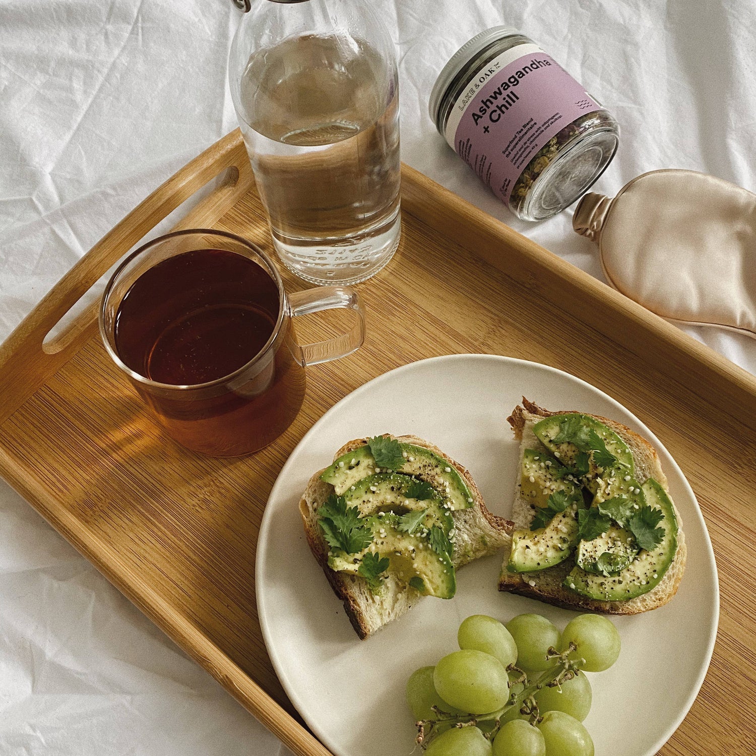 Wooden tray with a plate of toast, grapes, a glass of water, and a jar on a white surface.