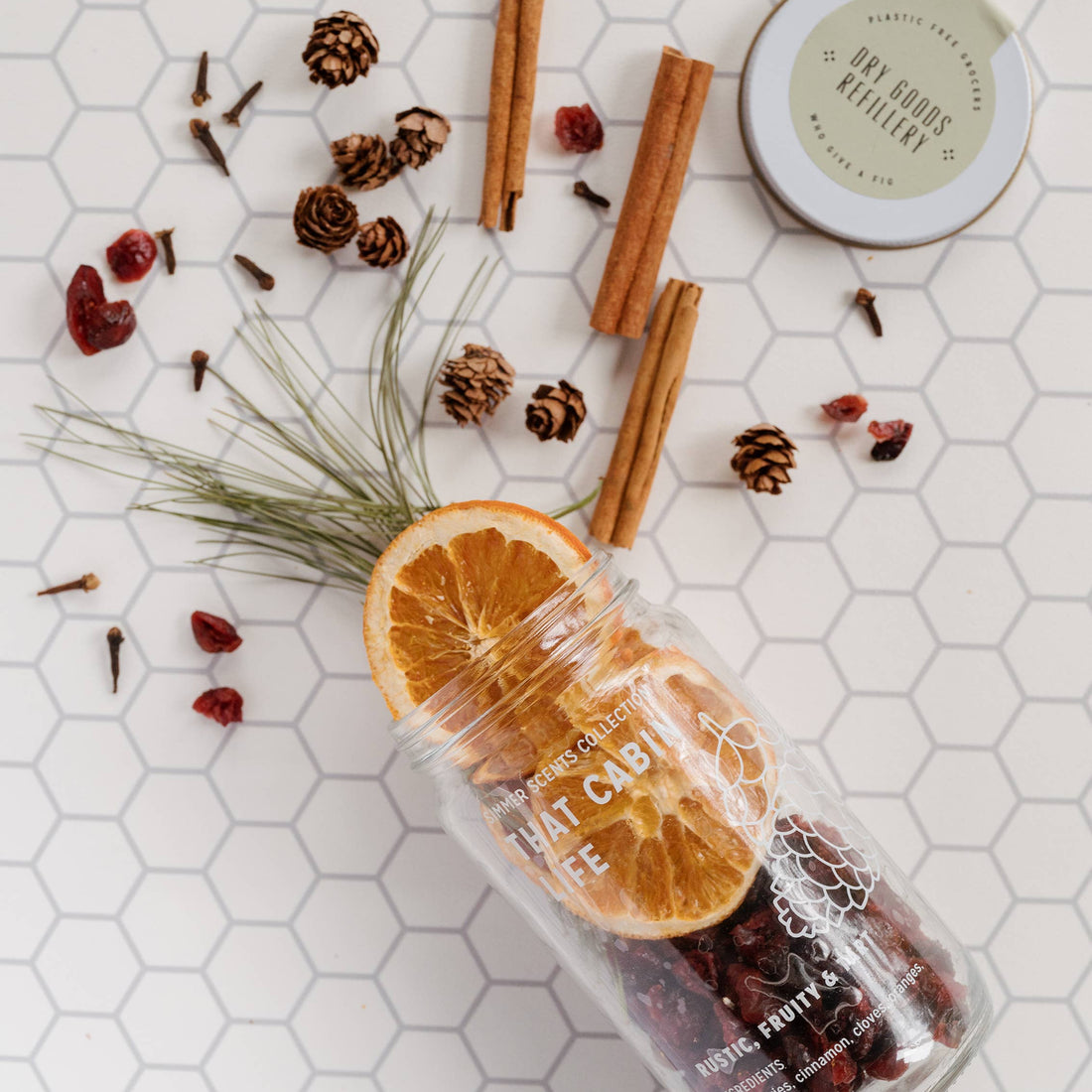 Spice mix in a glass bottle with orange slices, cinnamon sticks, and pine cones on a hexagonal tile background.
