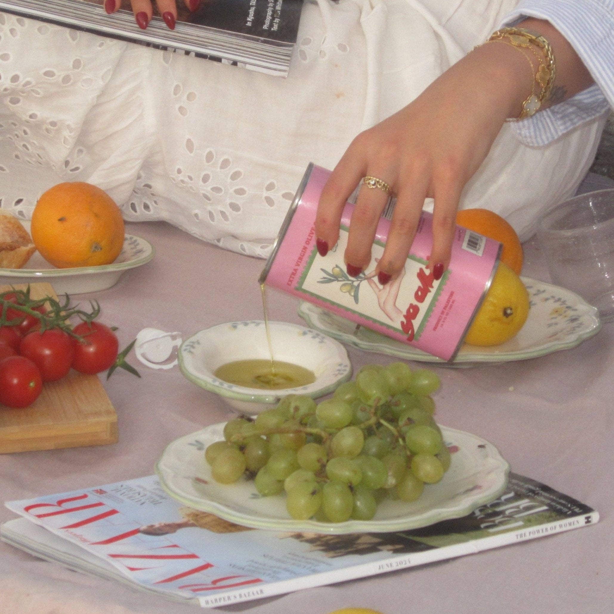 Person pouring olive oil into a glass with fruits and a magazine on a table