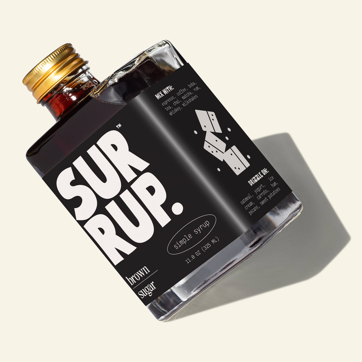 A bottle of SURRUP brand brown sugar simple syrup with a black label and gold cap, photographed on a light beige background with a soft shadow.