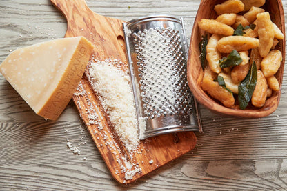 Grated Parmesan cheese on a wooden board with a cheese grater, a bowl of pasta, and a block of cheese.