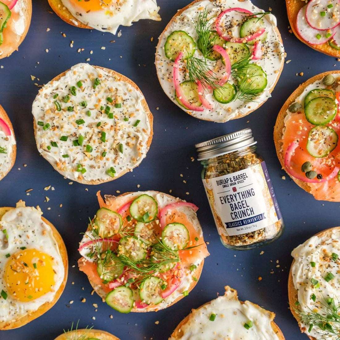 Assorted bagel slices with various toppings on a blue surface, featuring a jar of everything bagel crunch.