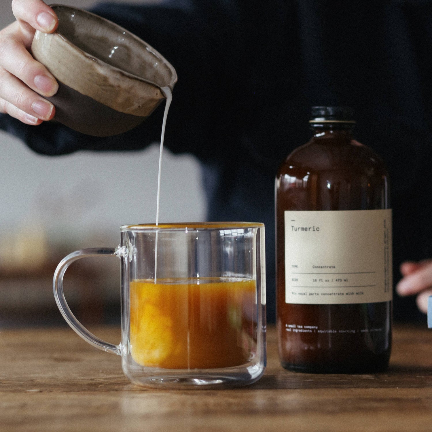 Person pouring liquid from a small bowl into a glass mug on a wooden surface with a bottle labeled &