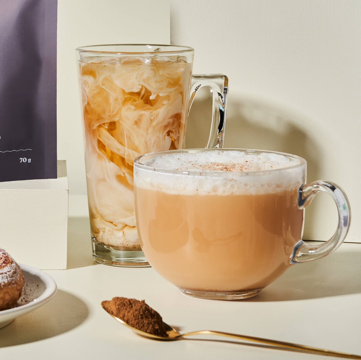 Two glass mugs with beverages, a spoon with cocoa powder, and a package on a light surface.