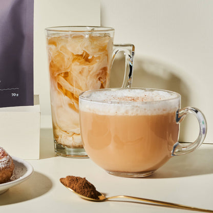 Two glass mugs with beverages, a spoon with cocoa powder, and a package on a light surface.