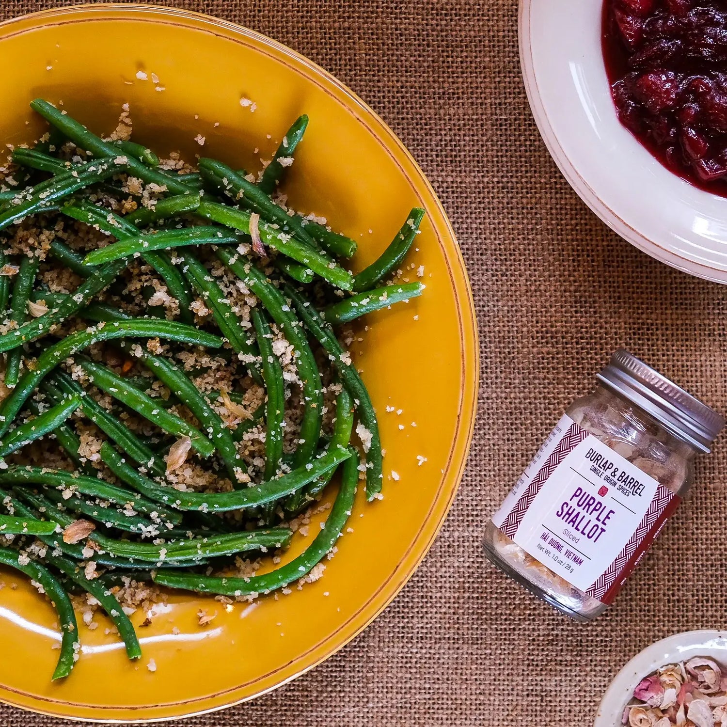 Yellow plate with green beans, jar of purple shallot seasoning, and bowl of cranberries on a brown textured surface.