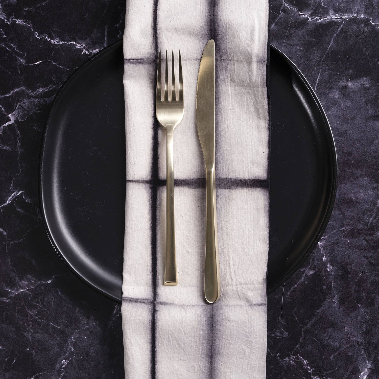Gold fork and knife on a folded white napkin with black lines, placed on a black plate against a dark marble background.