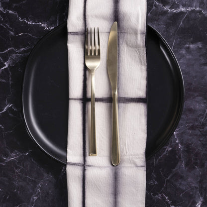 Gold fork and knife on a folded white napkin with black lines, placed on a black plate against a dark marble background.