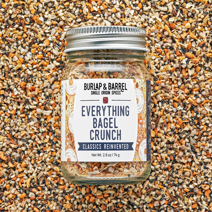 A glass jar of Burlap &amp; Barrel Everything Bagel Crunch spice mix, filled with ground spices and topped with a silver metal lid, set against a background of whole bagel and seed pieces.