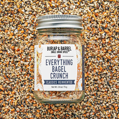 A glass jar of Burlap & Barrel Everything Bagel Crunch spice mix, filled with ground spices and topped with a silver metal lid, set against a background of whole bagel and seed pieces.