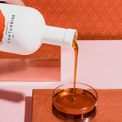 Bottle of Brightland oil being poured into a glass bowl against an orange textured background