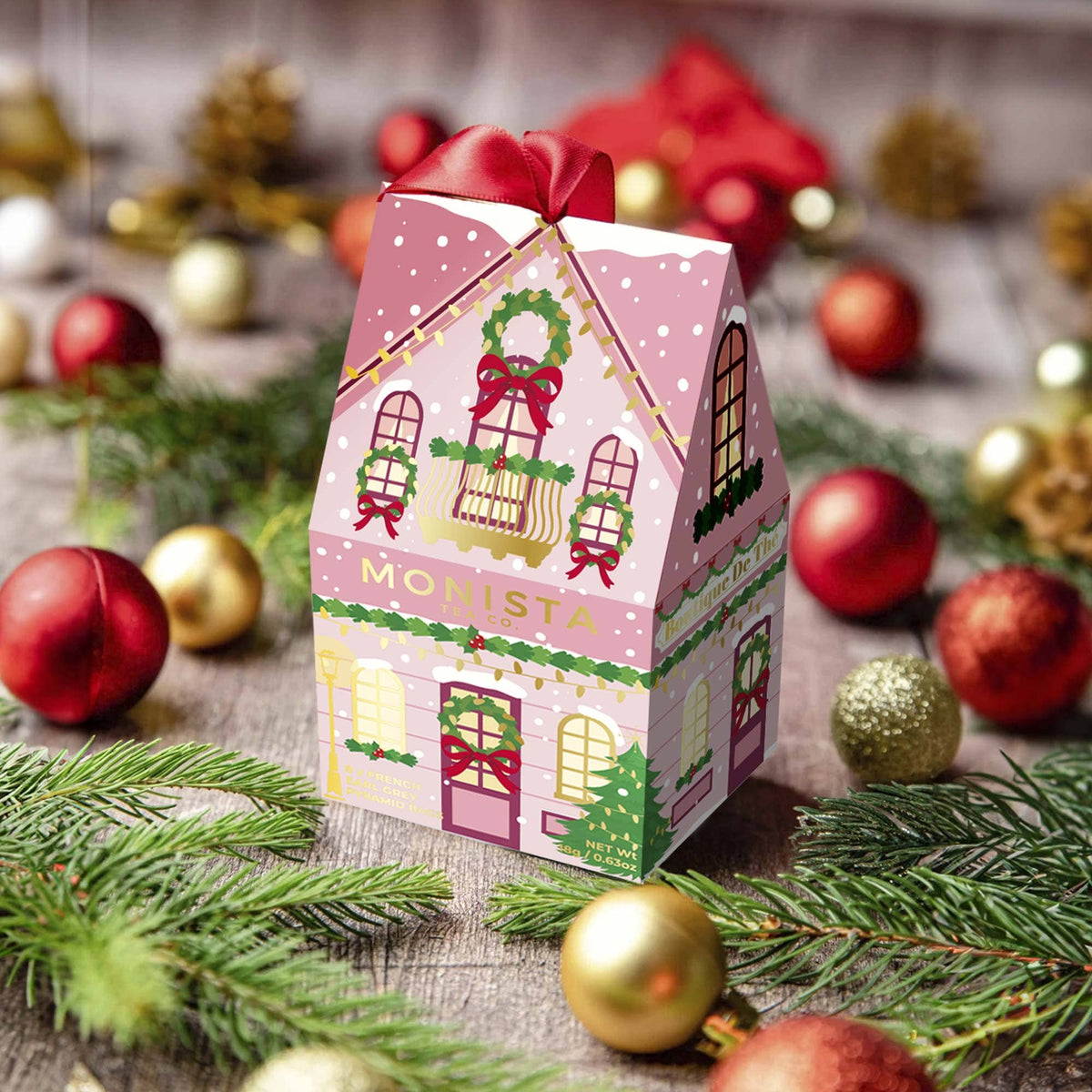 A pink Christmas-themed house-shaped candy box with a red bow and green wreath illustration, surrounded by holiday ornaments, pine branches, and a rustic wooden surface.