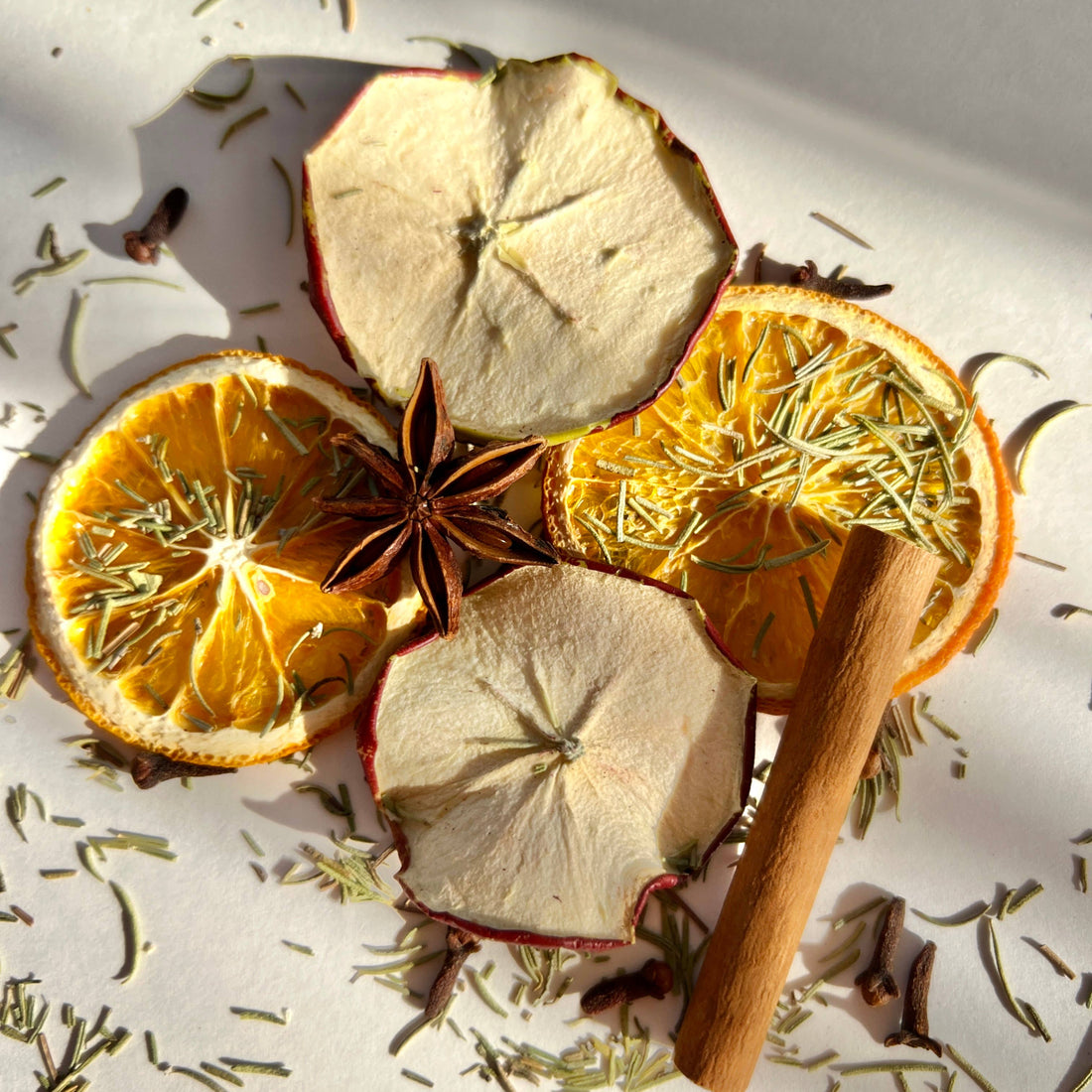 Dried oranges, apples, cinnamon stick, and star anise on a light surface with herbs.