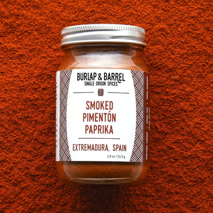 A glass jar of Burlap &amp; Barrel Smoked Pimenton Paprika spices, set against a textured orange-red background.
