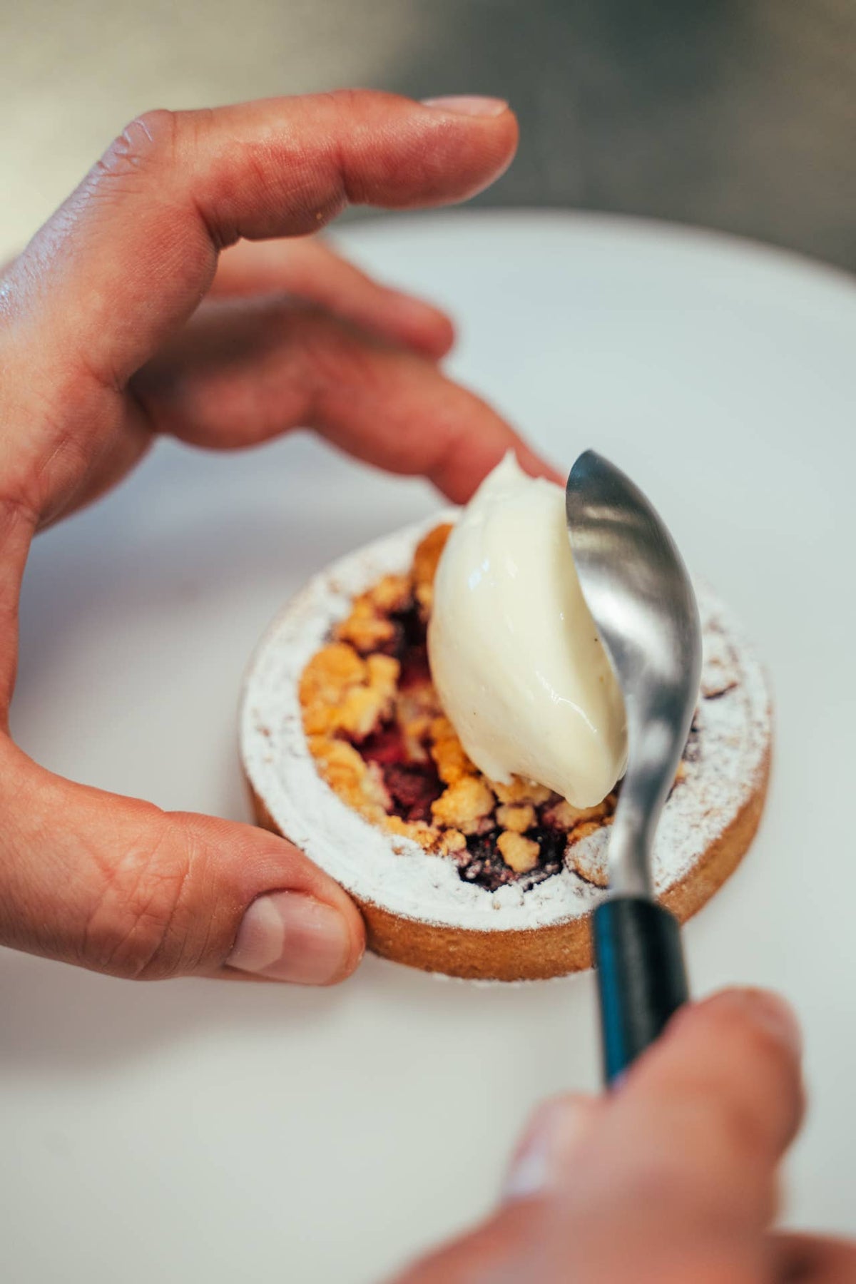 A close-up shot of a hand holding a small round pastry, while another hand uses a quenelle spoon to scoop cream onto it. The scene is shot from a high angle on a white plate or surface.