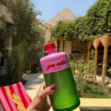 A hand holding a bottle of Ya Albi extra virgin olive oil with pink top and green body, set against a blurred outdoor patio background.