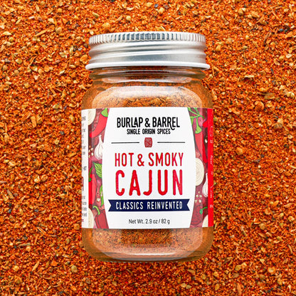A glass jar of Burlap &amp; Barrel Hot &amp; Smoky Cajun spices, filled with reddish-brown seasoning, set against a background of loose red and brown spices. 