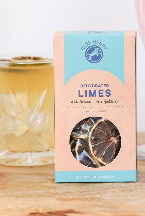 Blue Henry Dehydrated Limes dried fruit garnish in a box, shown on a wooden surface with a blurred cocktail in the background.