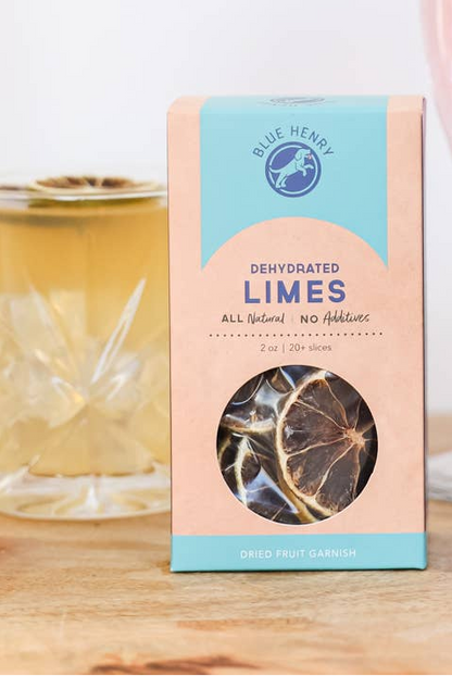 Blue Henry Dehydrated Limes dried fruit garnish in a box, shown on a wooden surface with a blurred cocktail in the background.