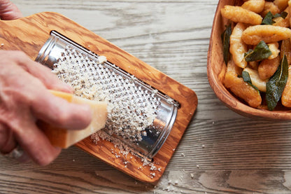 Hand grating cheese onto a wooden board with a bowl of gnocchi n the background.