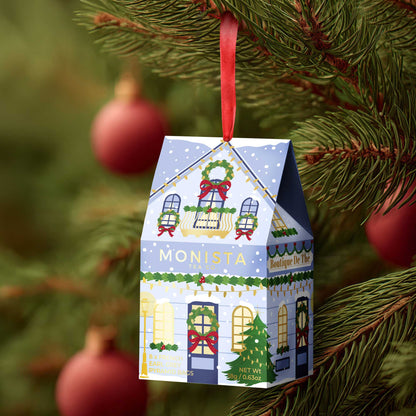 A small hanging Christmas ornament shaped like a house, printed with a festive village scene and the name MONISTA, suspended by a red ribbon from a pine branch with blurred red ornaments in the background.