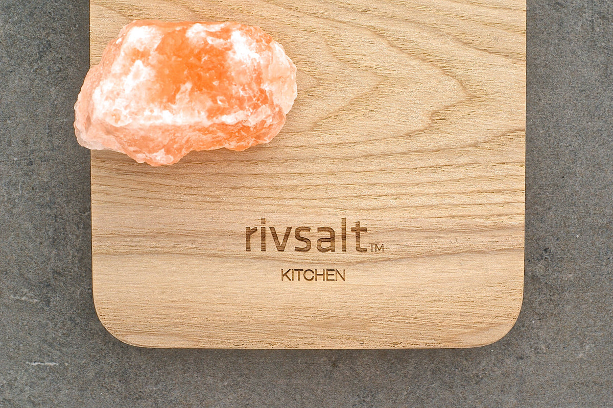 A pink salt crystal sits on a light wood cutting board engraved with the brand name &