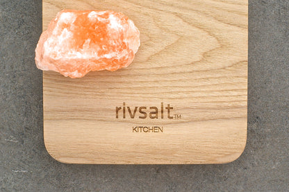A pink salt crystal sits on a light wood cutting board engraved with the brand name &