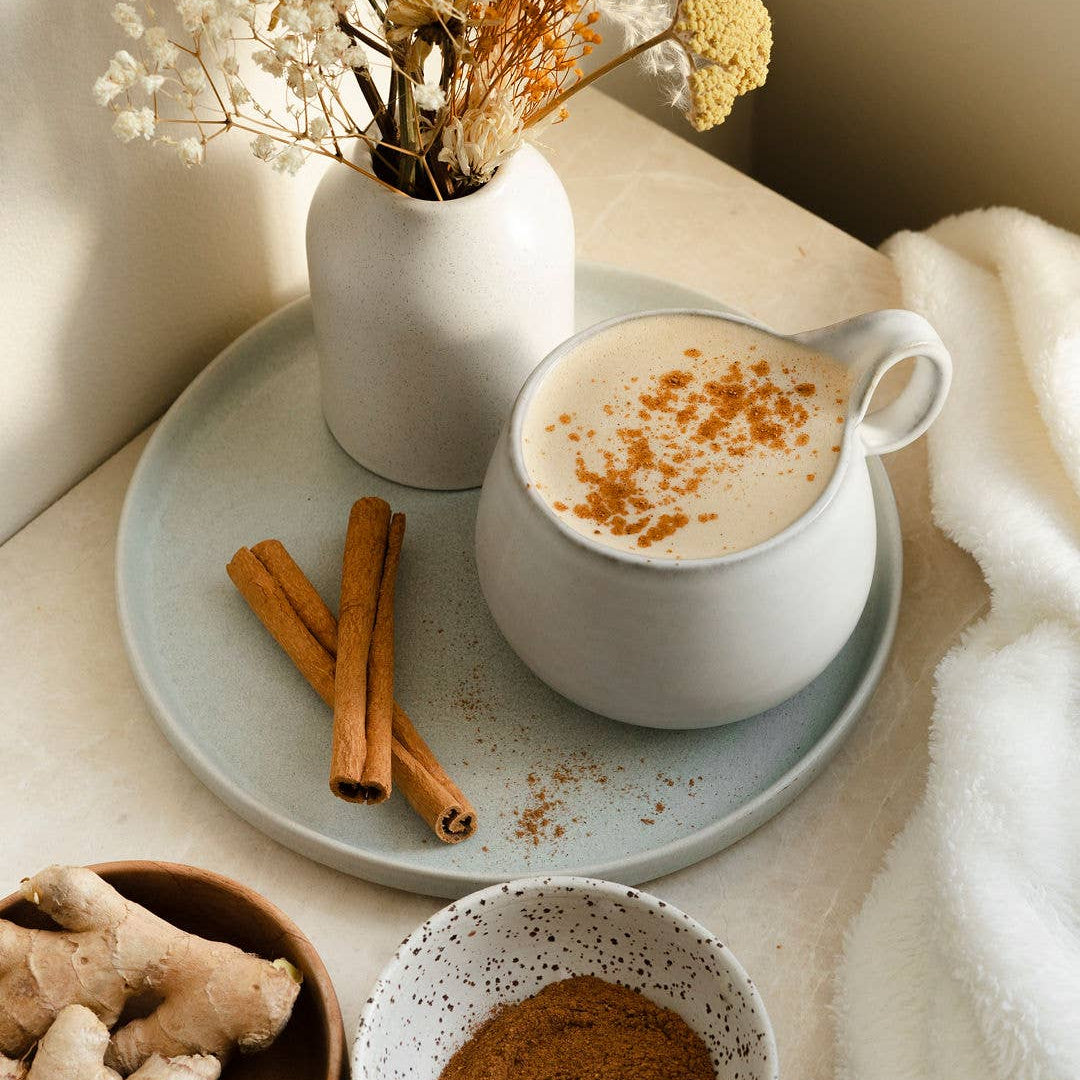 Cup of coffee with cinnamon on a tray, surrounded by ginger, cinnamon sticks, and a vase of flowers.