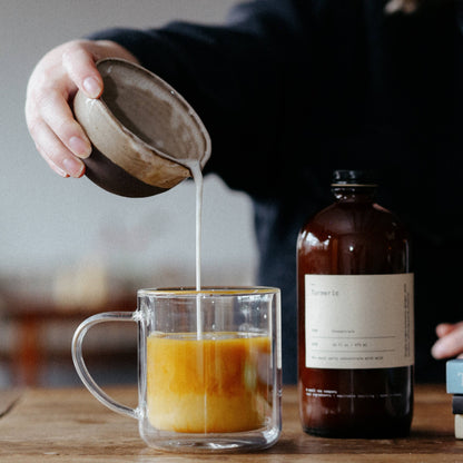 Person pouring liquid into a glass mug on a wooden table with a bottle in the background.