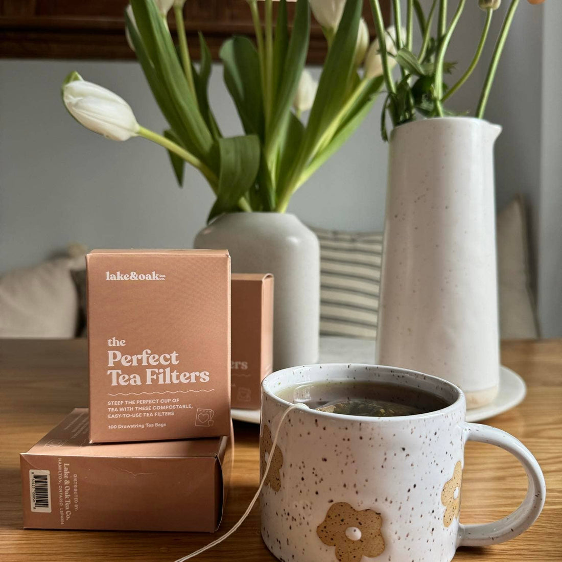 Tea cup with floral design, tea filters packaging, and flowers in vases on a wooden table.