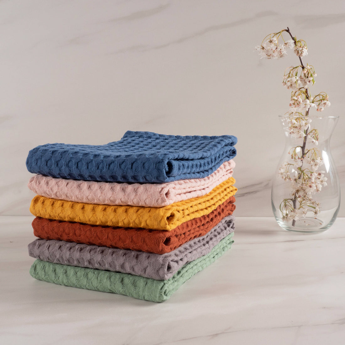 Stack of colorful textured towels on a light surface with a vase of flowers in the background.