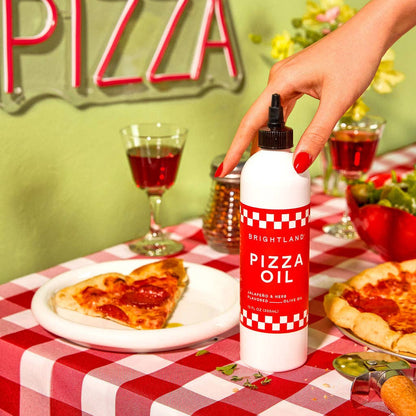 Person holding a bottle of pizza oil over a slice of pizza on a checkered tablecloth with a neon &
