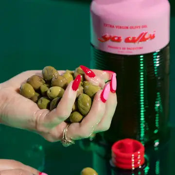 A hand with bright pink nails holds green olives in front of a pink and green bottle of Ya Albi Extra Virgin Olive Oil